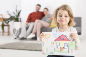 Photo of a smiling little girl holding a drawing with a house, with her parents sitting on a sofa in the blurry background